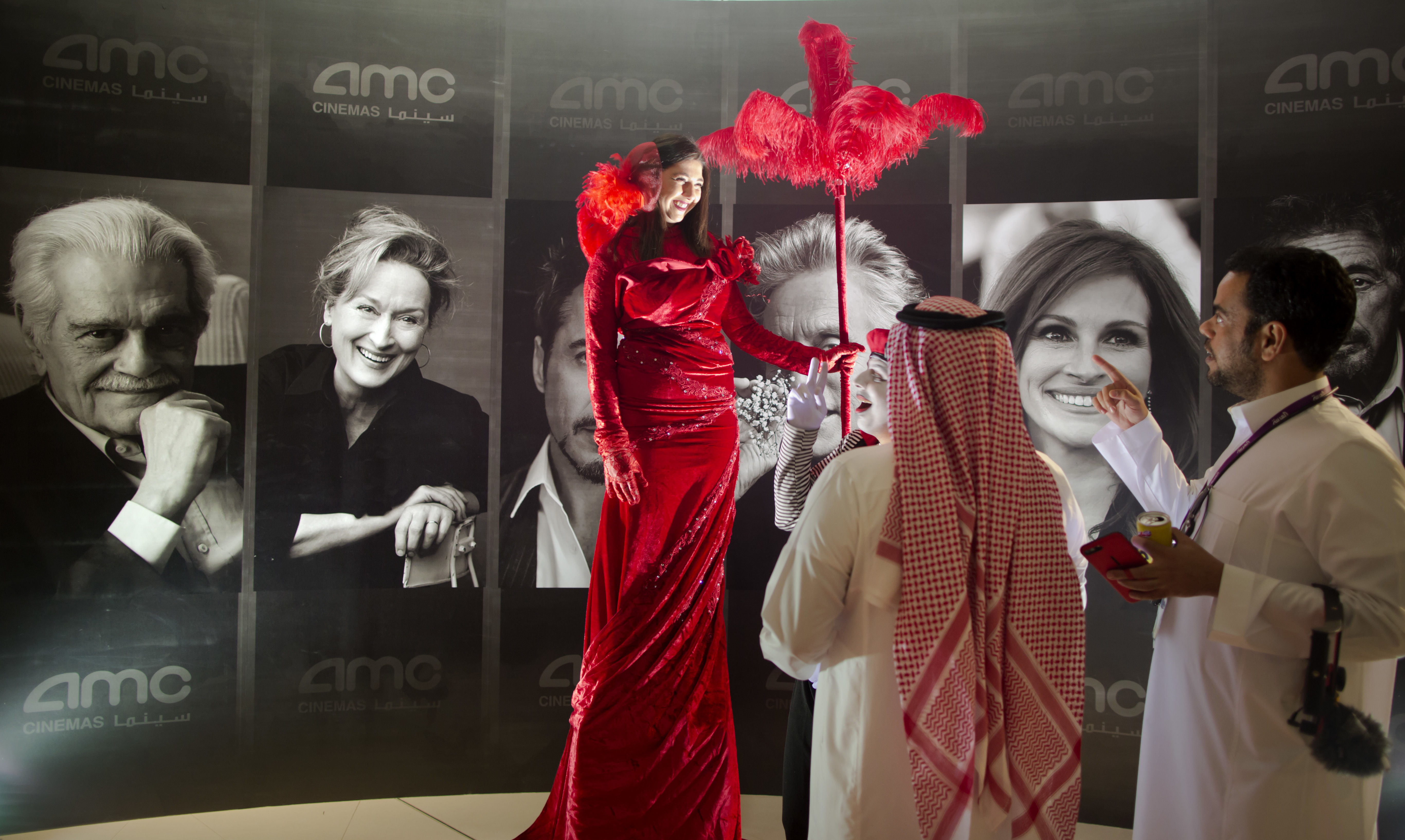 Visitors chat with an actress dress with red carpet colors in front of posters of Hollywood stars during invitation-only screening, at the King Abdullah Financial District Theater, in Riyadh, Saudi Arabia, Wednesday, April 18, 2018. Saudi Arabia held a private screening of the Hollywood blockbuster 