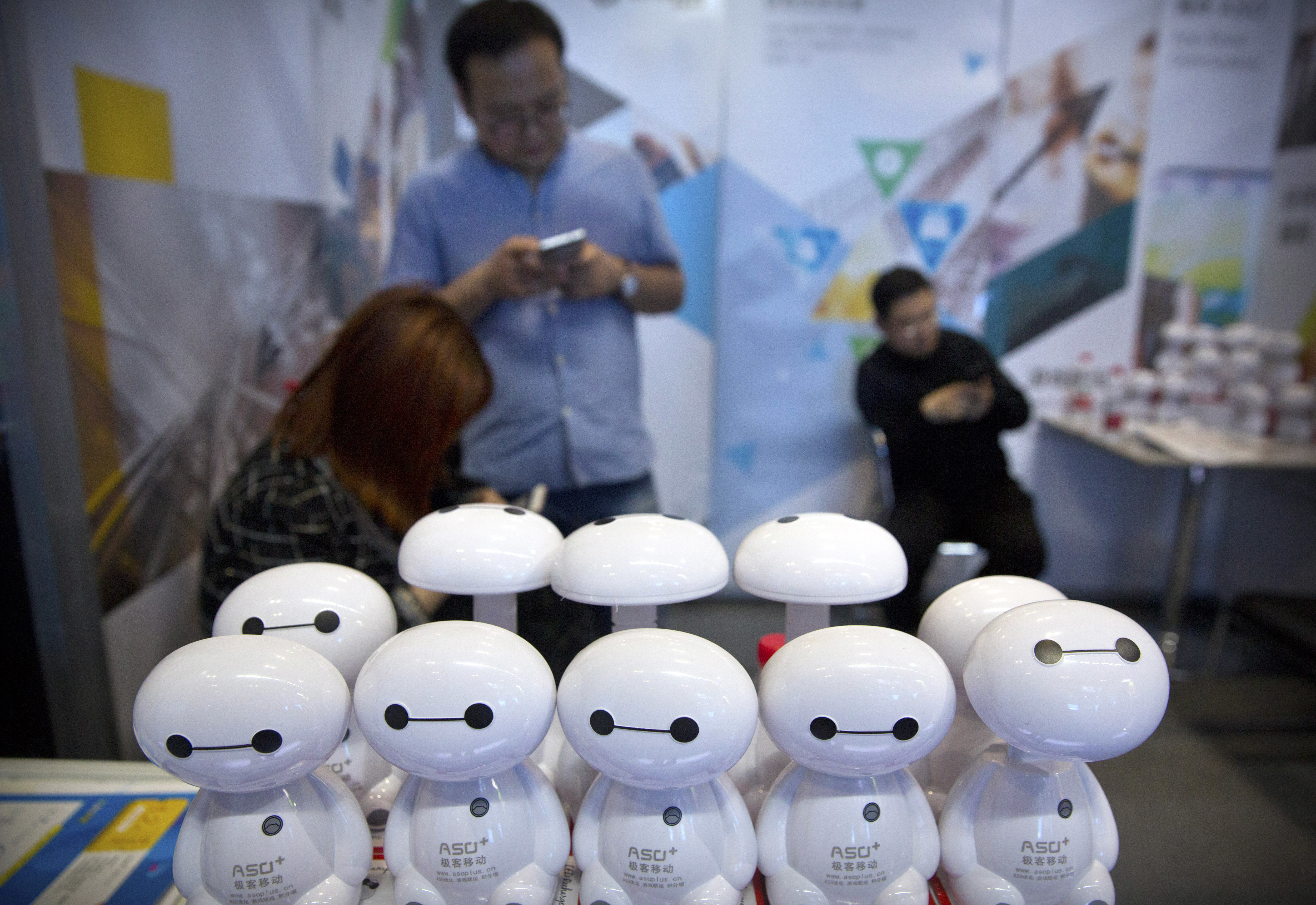 Staff members use their smartphones near a stack of plastic toy robots at a booth at the Global Mobile Internet Conference (GMIC) in Beijing, Thursday, April 26, 2018. The GMIC features current and future trends in the mobile Internet industry by some major foreign and Chinese internet companies. (AP Photo/Mark Schiefelbein)