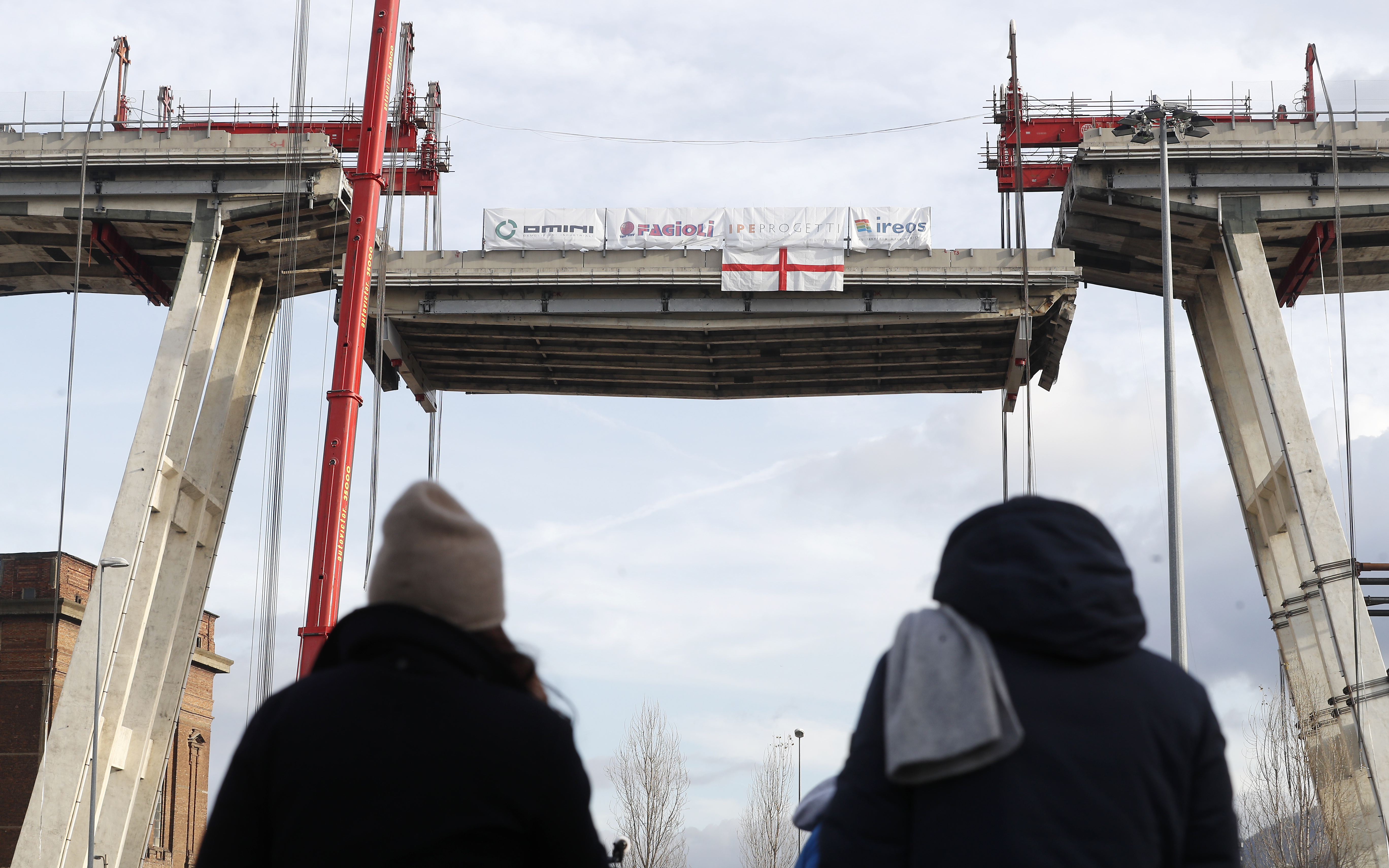 People look at cranes as they lower a section of the Morandi bridge in Genoa, Italy, Saturday, Feb. 9, 2019. Workers taking apart the remains of a bridge which collapsed in Aug. 2018 are set to remove a 40 meter beam, seen in between the red machinery. A large section of the bridge collapsed over an industrial area in the Italian city of Genova last summer during a sudden and violent storm, leaving vehicles crushed in rubble below and killing 43 people. (AP Photo/Antonio Calanni)
