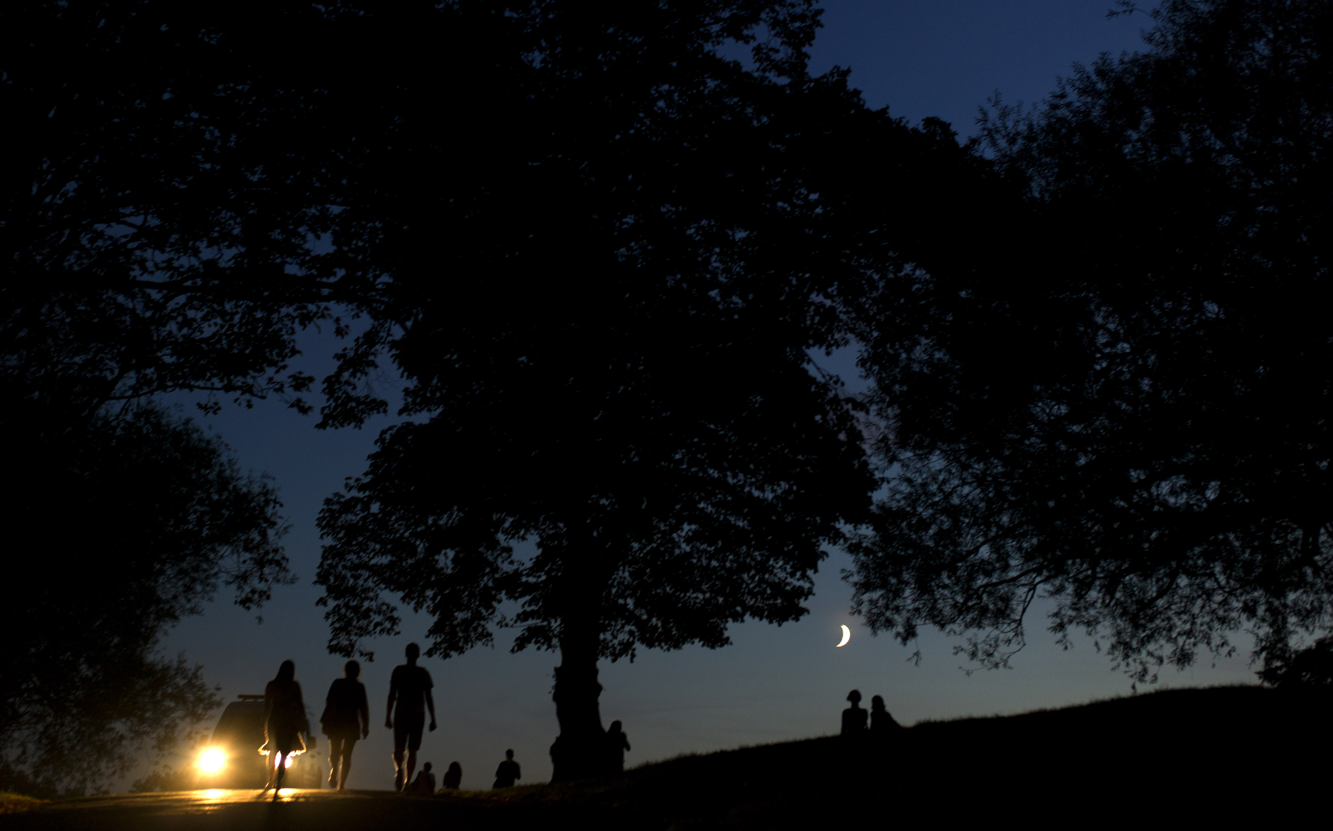 A police vehicle's headlights are seen as people are silhouetted in the fading light as the moon peers through the trees during unseasonal hot weather on Hampstead Heath in London, Saturday, Oct. 1, 2011.  Britain experienced the hottest October day on record Saturday, with the national weather service the Met Office saying the temperature reached 85.8 F (29.9 C) at Gravesend in southeast England.  That is the highest October temperature since records began a century ago, beating the previous high of 84.9 F (29.4 C) reached on Oct. 4, 1985.  (AP Photo/Matt Dunham)