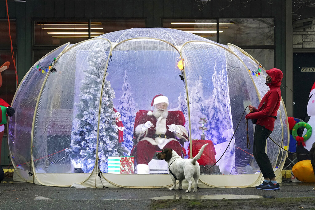 FILE - In this Dec. 8, 2020, file photo, Santa, portrayed by Dan Kemmis, laughs as he talks to Kristin Laidre as she walks her dog, Scooby, a Bassett Hound mix, as he sits inside a protective bubble in Seattle's Greenwood neighborhood. All most people wanted for Christmas after this year of pandemic was some cheer and togetherness. Instead many are heading into a season of isolation, grieving lost loved ones, experiencing uncertainty about their jobs or confronting the fear of a potentially more contagious variant of the coronavirus.(AP Photo/Ted S. Warren, File)