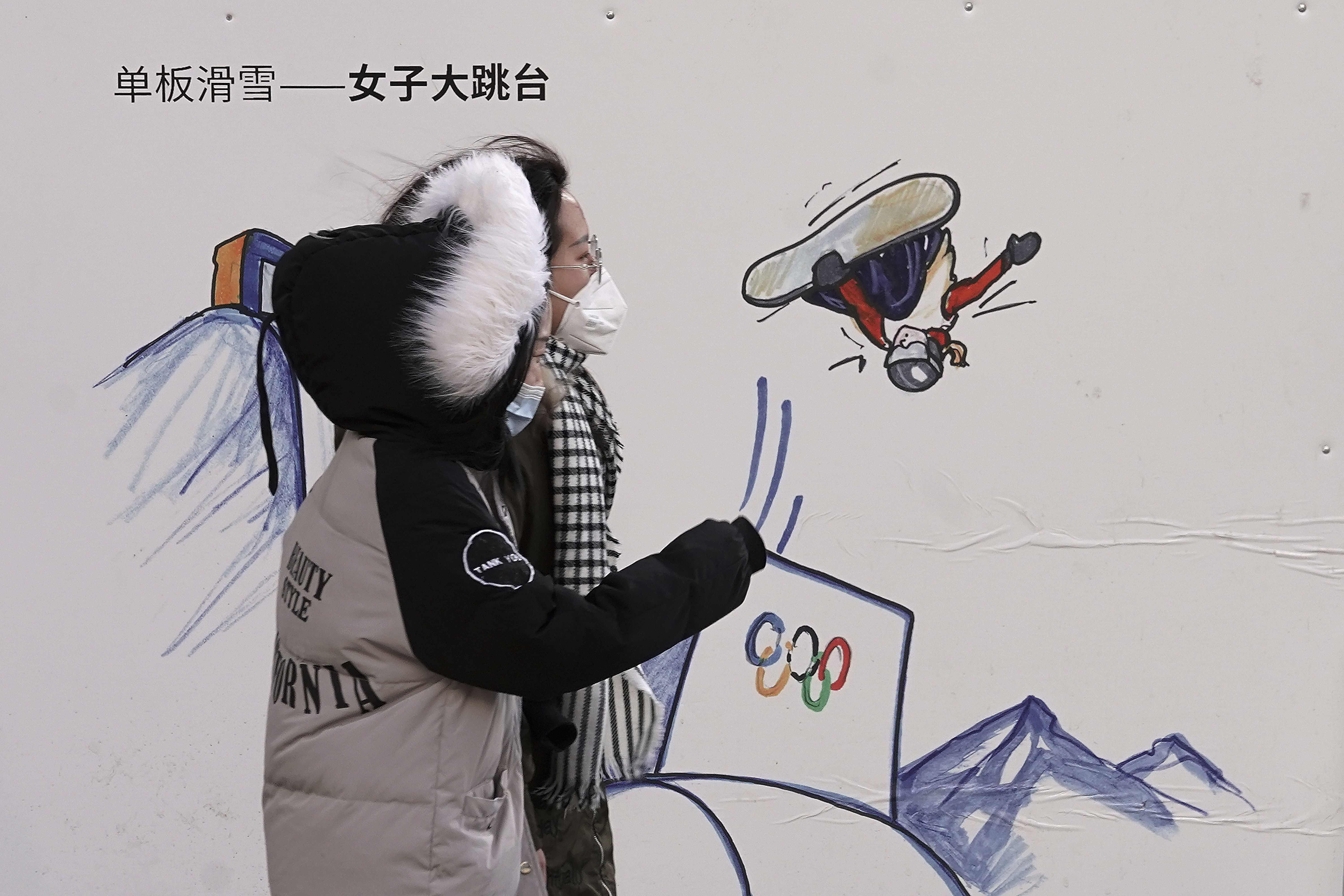 Women wearing face masks to help curb the spread of the coronavirus walk by a board depicting a Winter Olympics snowboard event at a shopping mall in Beijing, Wednesday, Jan. 6, 2021. China's Hebei province is enforcing stricter control measures following a further rise in coronavirus cases in the province, which is adjacent to the capital Beijing and is due to host events for next year's Winter Olympics. (AP Photo/Andy Wong)