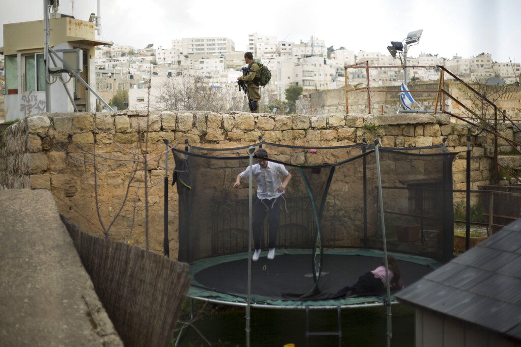 FILE - In this March 7, 2019, file photo, settlers jump on a trampoline as an Israeli solider stands guard in the Israeli controlled part of the West Bank city of Hebron. Israel's premier human rights group has begun describing both Israel and its control of the Palestinian territories as a single 