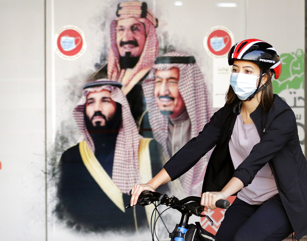 Sawsan Abdel Fattah, a Saudi cyclist member of women's brave cyclist team, rides her bicycle in front of a banner showing Saudi King Salman, right, his Crown Prince Mohammed bin Salman, left, and Saudi Arabia's founder late King Abdul Aziz Al Saud in Jiddah, Saudi Arabia Saturday, March 6, 2021. The brave cyclist team, which was formed in 2019 aiming to normalize the sport for women, organize a tour cycling ahead of International Women's Day. (AP Photo/Amr Nabil)