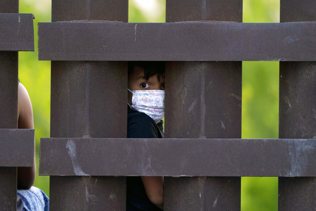 FILE - In this March 21, 2021, file photo, a migrant child peeks through the U.S.-Mexico border metal wall as a group of migrants is processed and taken into custody while trying to sneak across the border in Abram-Perezville, Texas. For the third time in seven years, U.S. officials are scrambling to handle a dramatic spike in children crossing the U.S.-Mexico border alone, leading to a massive expansion in emergency facilities to house them as more kids arrive than are being released to close relatives in the United States. (AP Photo/Julio Cortez, File)