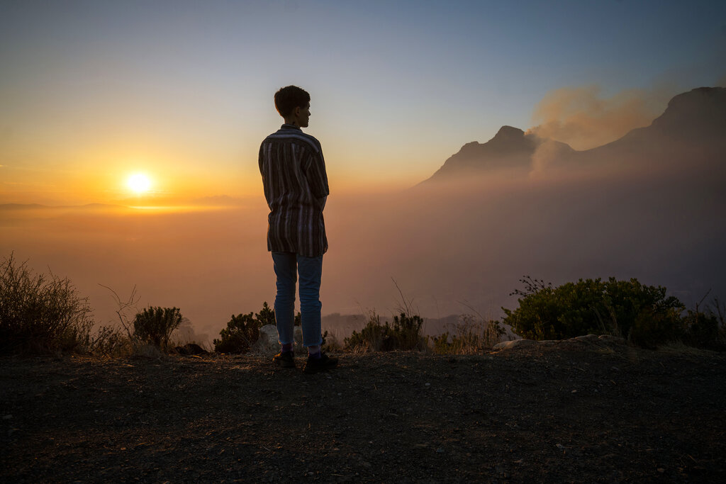 A Capetonian watches the sun rise from the top of Signal Hill as smoke engulfs the city of Cape Town, South Africa, Tuesday April 20, 2021. A massive fire spreading on the slopes of the city's famed Table Mountain, at right, is kept under control as firemen and helicopters take advantage of the low winds to contain the blaze. (AP Photo/Jerome Delay)