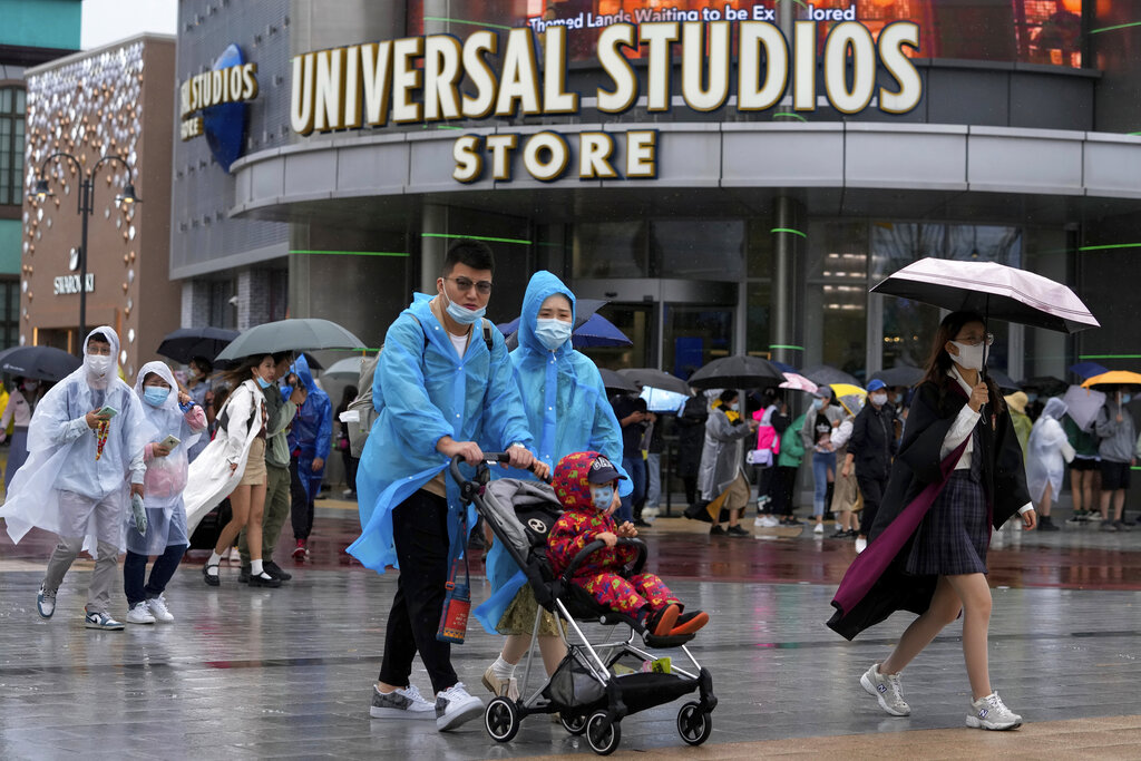 A family wearing raincoats and face masks to help protect from the coronavirus walk by masked visitors lining up to enter a Universal Studios store as they visit Universal Studios Beijing in Beijing, Monday, Sept. 20, 2021. Thousands of people brave the rain visit to the newest location of the global brand of theme parks which officially opens on Monday. (AP Photo/Andy Wong)