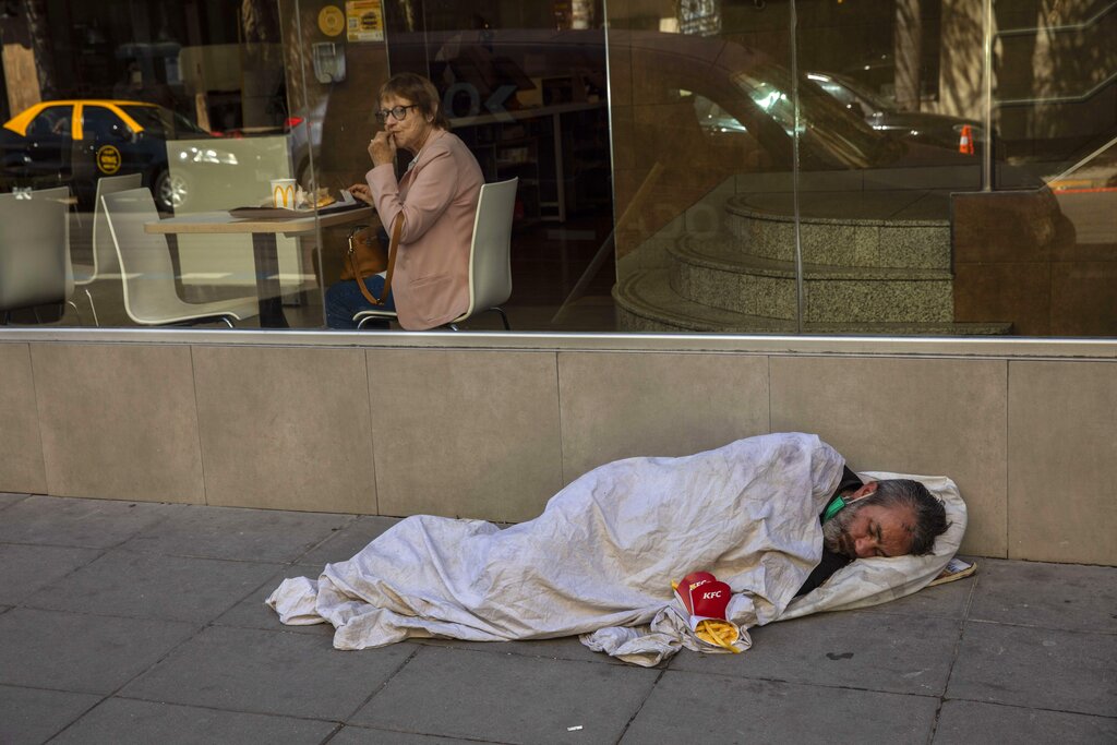 A woman eats lunch in a fast food restaurant while a homeless man sleeps on a sidewalk in the streets of Buenos Aires, Argentina, Thursday, Nov. 11, 2021. In the midst of a severe economic crisis, legislative elections are scheduled for Nov. 14. (AP Photo/Rodrigo Abd)
