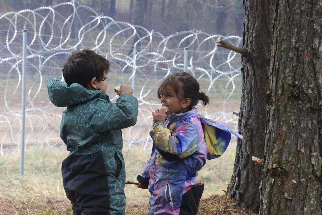 Migrant children eat near the barbed wire fence as migrants gather at the Belarus-Poland border near Grodno, Belarus, Saturday, Nov. 13, 2021. A large number of migrants are in a makeshift camp on the Belarusian side of the border in frigid conditions. Belarusian state news agency Belta reported that Lukashenko on Saturday ordered the military to set up tents at the border where food and other humanitarian aid can be gathered and distributed to the migrants. (Leonid Shcheglov/BelTA pool photo via AP)