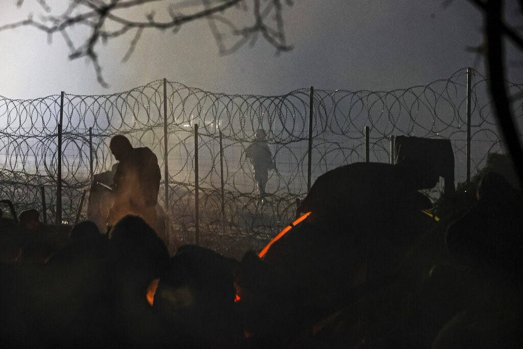 A Polish border guard patrols near a barbed wire fence as migrants warm themselves near fire gathering at the Belarus-Poland border near Grodno, Belarus, late Sunday, Nov. 14, 2021.European Union foreign ministers are expected Monday to decide to expand sanctions against Belarus to include airlines, travel agents and individuals alleged to be helping to lure migrants to Europe as part of a 