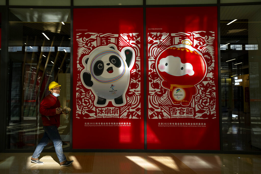 A worker wearing a face mask walks past posters showing the Olympic mascot Bing Dwen Dwen and Paralympic mascot Shuey Rhon Rhon at a commercial plaza at the Winter Olympic Village in Beijing, Friday, Dec. 24, 2021. Organizers on Friday gave the media a look at parts of the athletes' Olympic Village for the 2022 Winter Olympics and Paralympics, which will be held beginning in February. (AP Photo/Mark Schiefelbein)