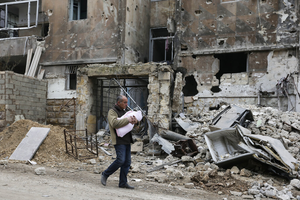 FILE - In this Jan. 20, 2017 photo, a man carries a baby as he walks past rubble in the once rebel-held Salaheddine neighborhood of eastern Aleppo, Syria. Tuesday, March 15, 2022 marks the 11th anniversary of Syria's revolution-turned-civil war. This year, many survivors are watching in shock as Ukrainians face the same horrors they did. (AP Photo/Hassan Ammar, File)