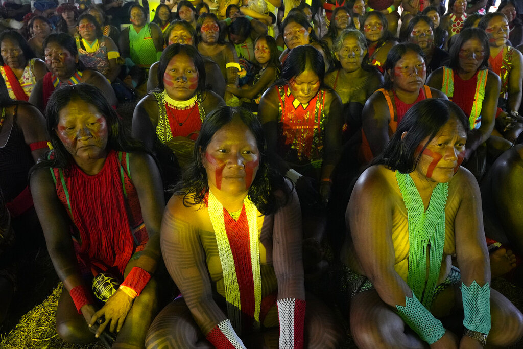 Indigenous women are illuminated by stage lighting as they listen to speakers during a ceremony called the Meeting of the First Peoples during the 18th annual Free Land Indigenous Camp in Brasilia, Brazil, Friday, April 8, 2022. The 10-day event aims to show the unity of Brazil's Indigenous peoples in their fight for the demarcation of their lands and their rights. (AP Photo/Eraldo Peres)