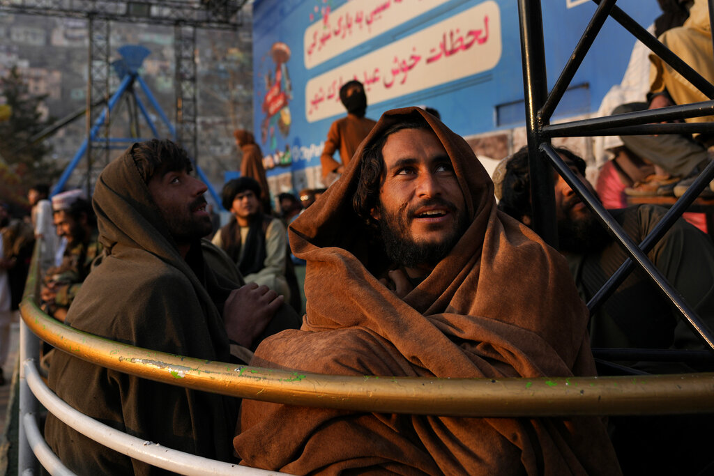 Afghan men cover themselves with blankets to warm from the cold weather, as they watch people enjoying at an amusement park, in Kabul, Afghanistan, Friday, Feb. 18, 2022. (AP Photo/Hussein Malla)
