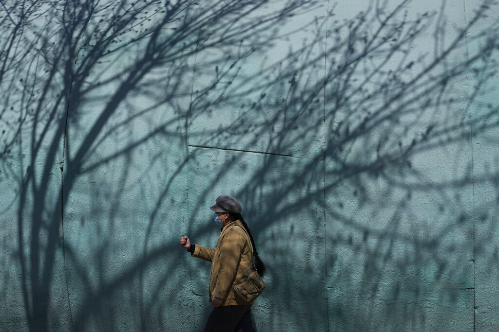A resident wearing a mask walks past shadows of a tree in a park on Thursday, March 31, 2022, in Beijing. China's case numbers in its latest infection surge are low compared with other major countries. But the ruling Communist Party is enforcing a 
