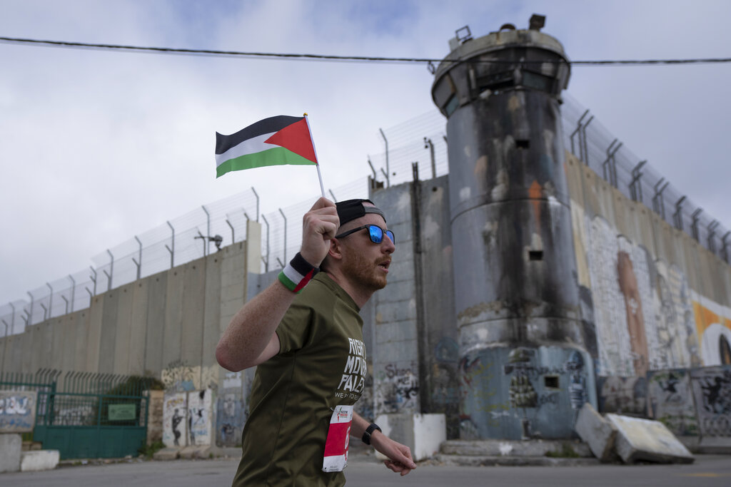 A runner carries a Palestinian flag and passes by the Israeli separation barrier during the Palestine eighth international Marathon, in the West Bank city of Bethlehem, Friday, March 18, 2022. After two years of cancellation due to the COVID-19 pandemic, the four tracks marathon kicked off today with the participation of over 10000 Palestinian and international athletes from eighty countries, with a 50 percent women participants, the marathon organizers said. (AP Photo/Nasser Nasser)