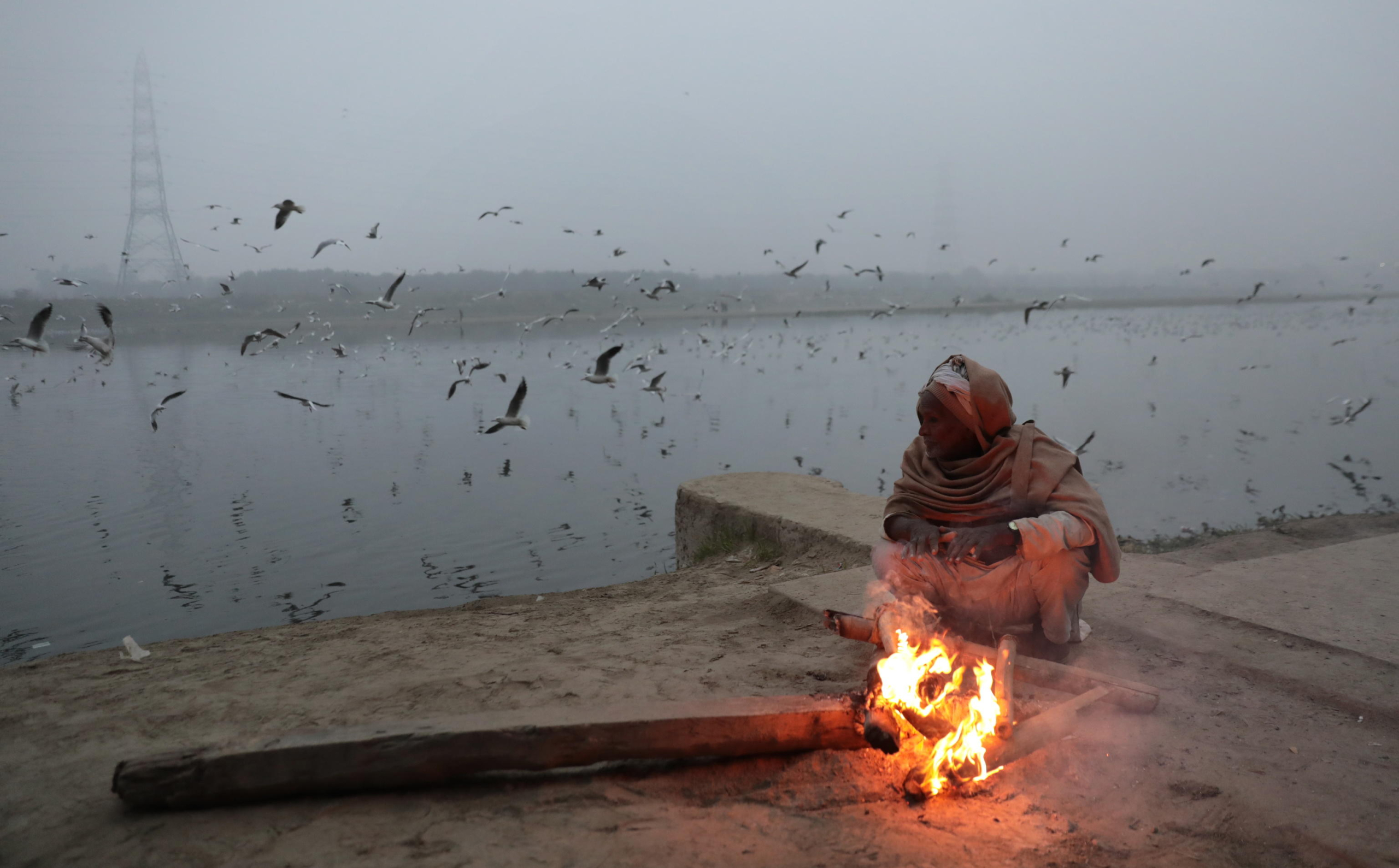 epa11058149 An Indian tries to warm himself near a fire as migratory birds are seen behind him on a foggy, cold winter morning on the banks of the Yamuna river in New Delhi, India, 05 January 2024. Winter takes a heavy toll each year around northern India, as poverty forces many homeless people to live outdoors. Temperatures in the northern Indian regions continue to decrease. Migratory birds, including seagulls, arrive at Delhis Yamuna bank during the onset of winter season as they migrate through Delhi making a temporary home in Yamuna river and sometime fed by devotees who arrive to bathe in the river in the morning time.  EPA/RAJAT GUPTA