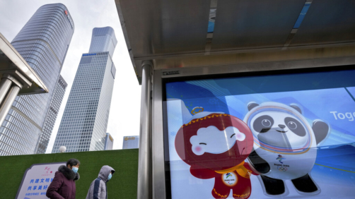 Commuters wearing face masks to help protect from the coronavirus walk by a bus stand displaying a poster showing mascot for the Beijing Winter Olympics at the central business district in Beijing, Wednesday, Jan. 26, 2022. (AP Photo/Andy Wong)